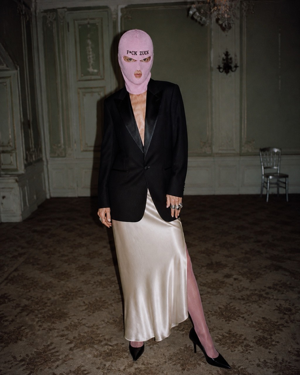 Model in the blush FACECRIME balaclava wearing a tuxedo jacket and satin skirt in a faded ballroom.